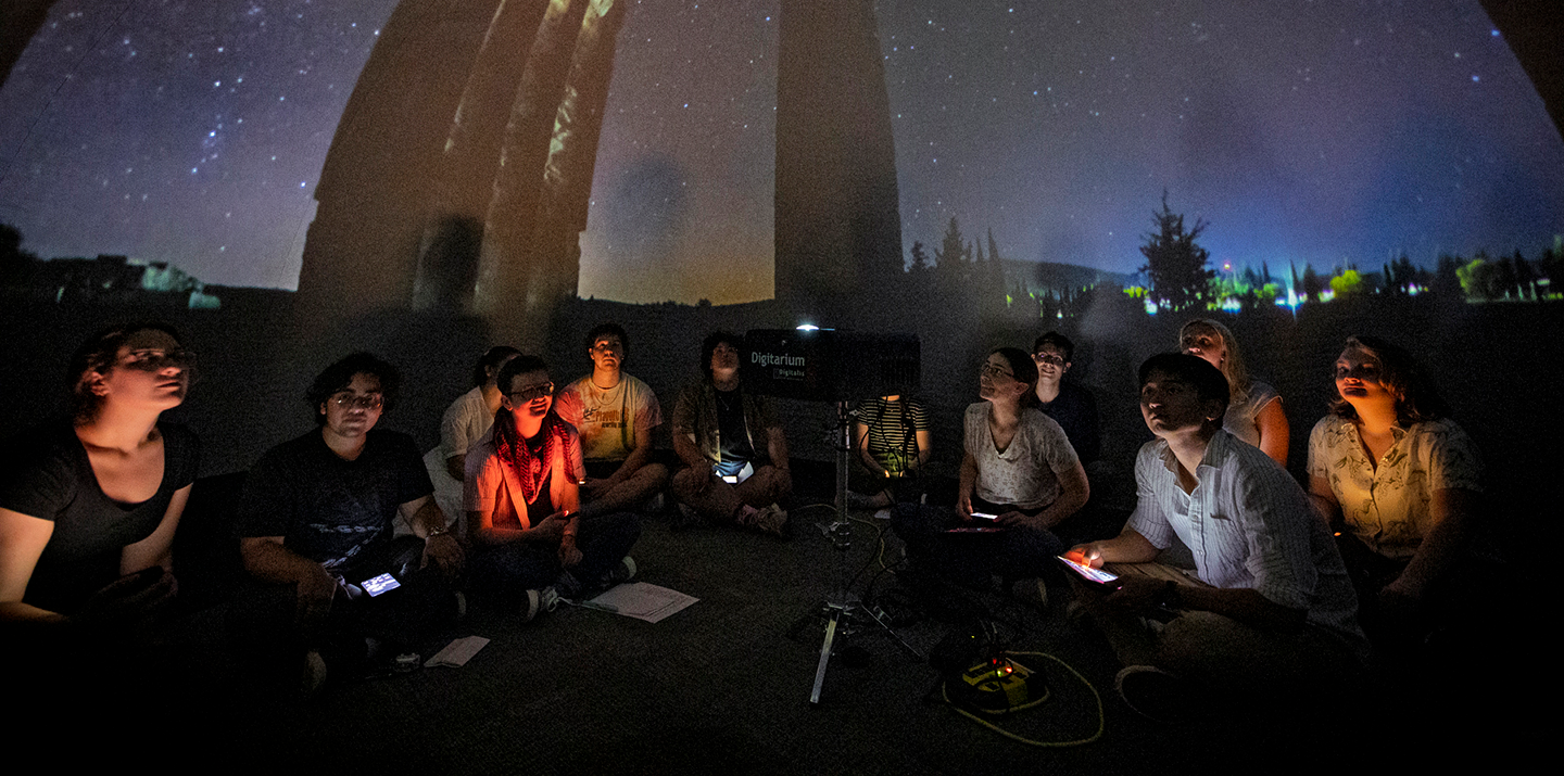 Students sit in a dark planetarium observing stars projected on the ceiling.