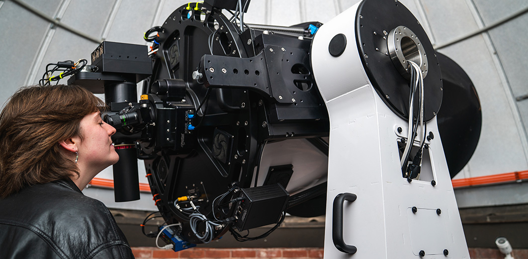 A student peers through the 24-inch PlaneWave telescope, surrounded by wires and observing equipment.