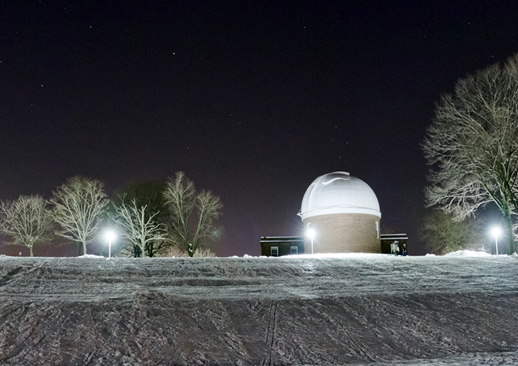Van Vleck Observatory dome at night, surrounded by snow and lit trees under a starry sky.