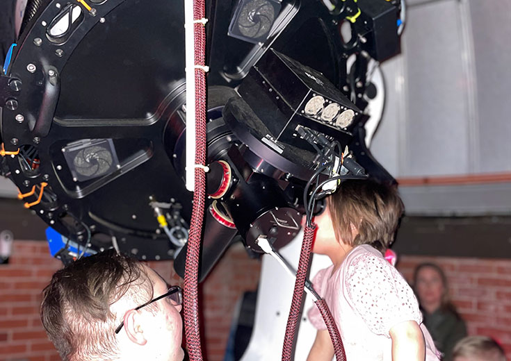 A young child looks through a large telescope while adults assist during a kids' night.