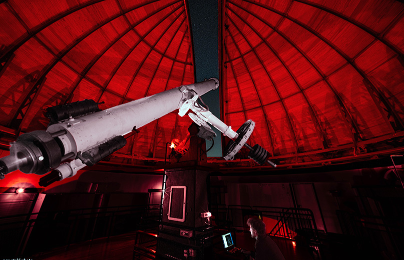 The Alvan Clark Refractor glows under red lights inside the observatory dome, with a starry sky above.