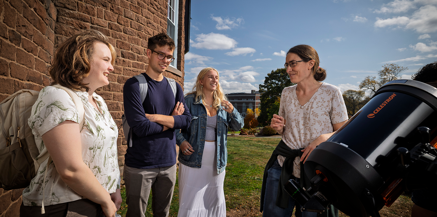 A professor and students chat outside by a telescope, smiling in the sun near the observatory.