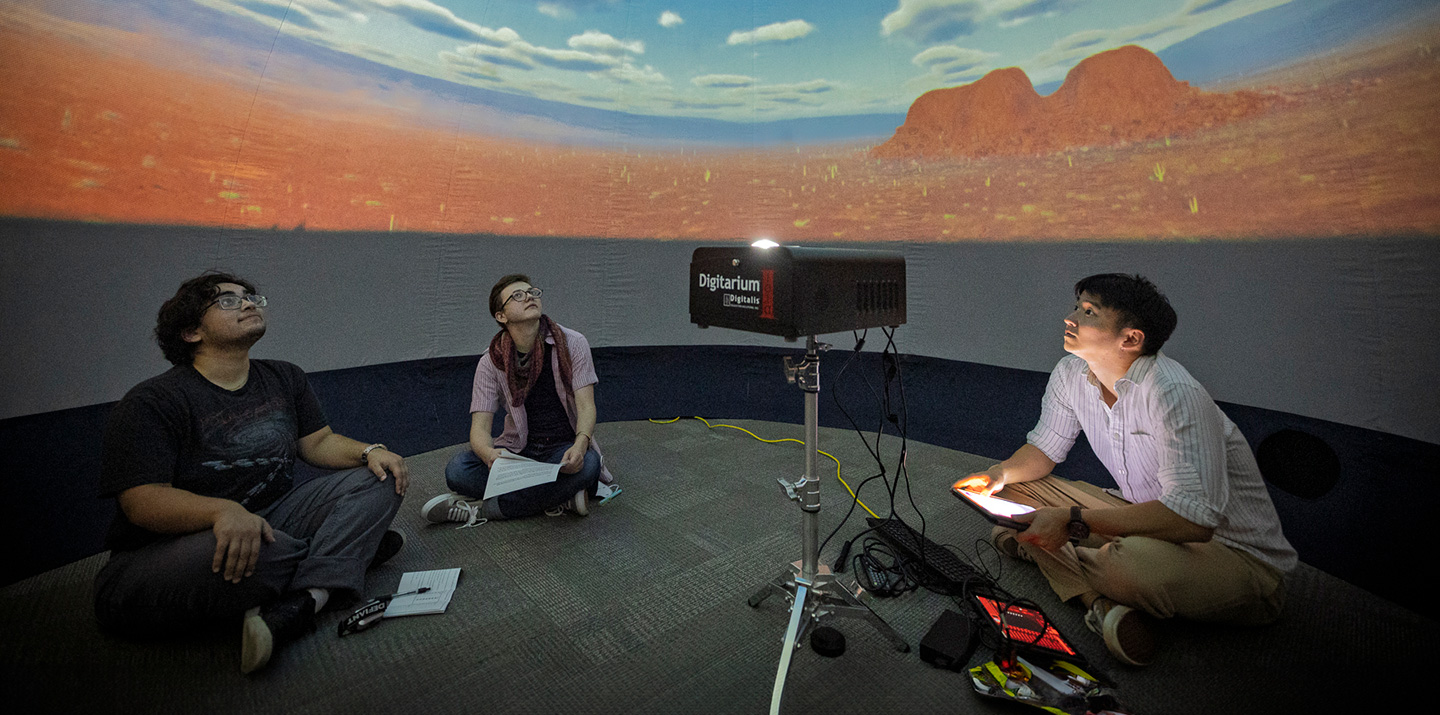Three students sitting cross-legged inside a planetarium dome, watching an immersive space projection above them.