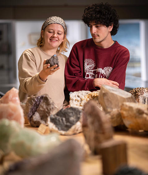 Two students indoors examining a rock or meteorite specimen, engaged in conversation, with shelves of books behind them