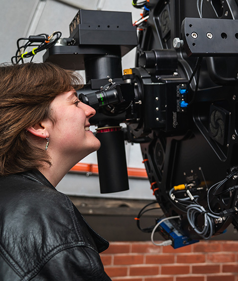 A student peers into a complex telescope mounted indoors, smiling as she adjusts the eyepiece, surrounded by scientific equipment.