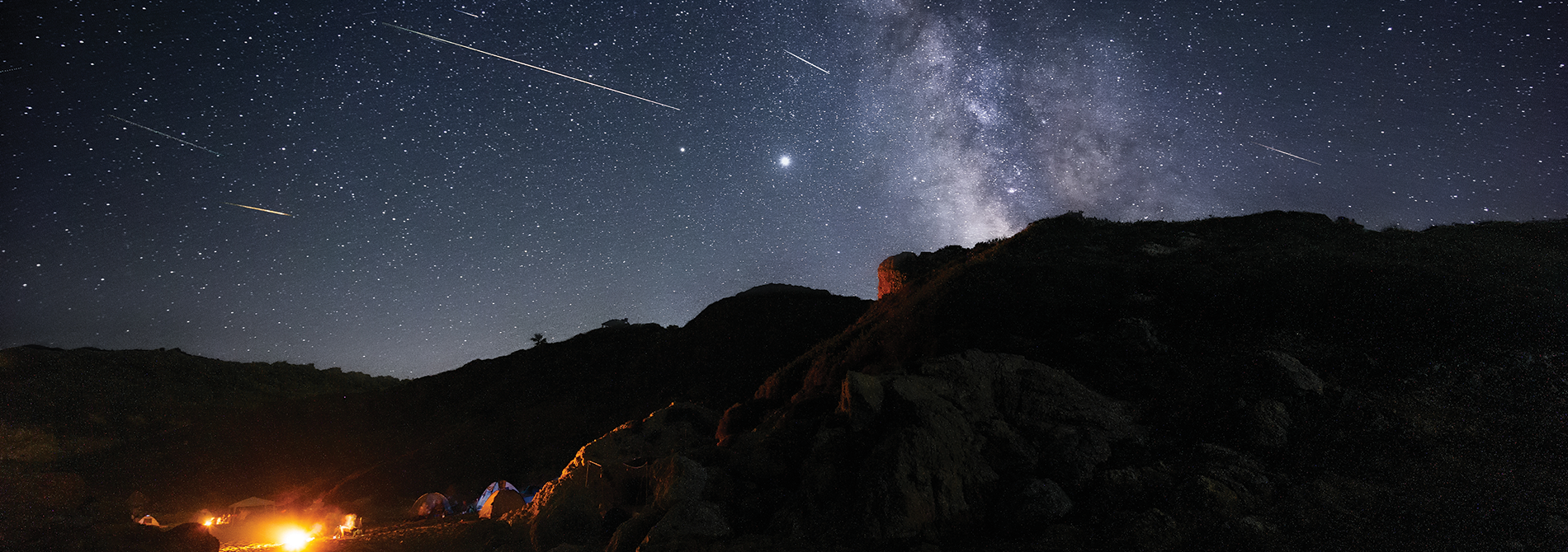 A mountain landscape under a star-filled sky with streaking meteors and a view of the Milky Way.