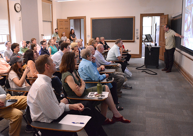 A professor gestures to a screen during a lecture as a full classroom of students and community members watch attentively.
